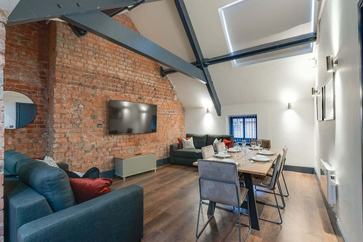 A modern living room with a brick wall, a flat-screen TV, a dining area with a table set for four, and a cozy seating area with a sofa and chairs.