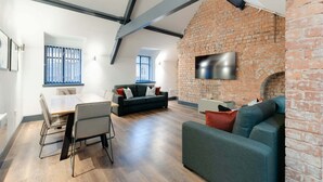 A modern living space with a brick wall, a flat-screen TV mounted above it, a dining area with a table and chairs, and a sofa. - Harrisons Aparthotel (Liverpool)