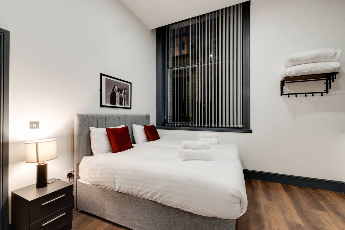 A neatly made bed with white linens and red pillows, a bedside lamp, a framed photo on the wall, and a window with blinds.