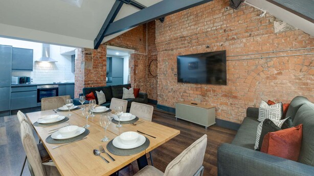 A modern kitchen with a brick wall, a dining area with a wooden table set for six, and a grey sofa. - Harrisons Aparthotel (Liverpool)
