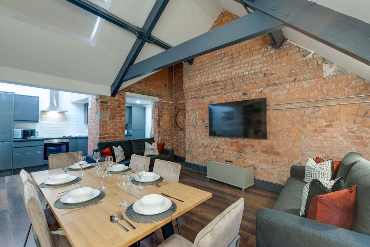 A modern kitchen with a brick wall, a dining area with a wooden table set for six, and a grey sofa.