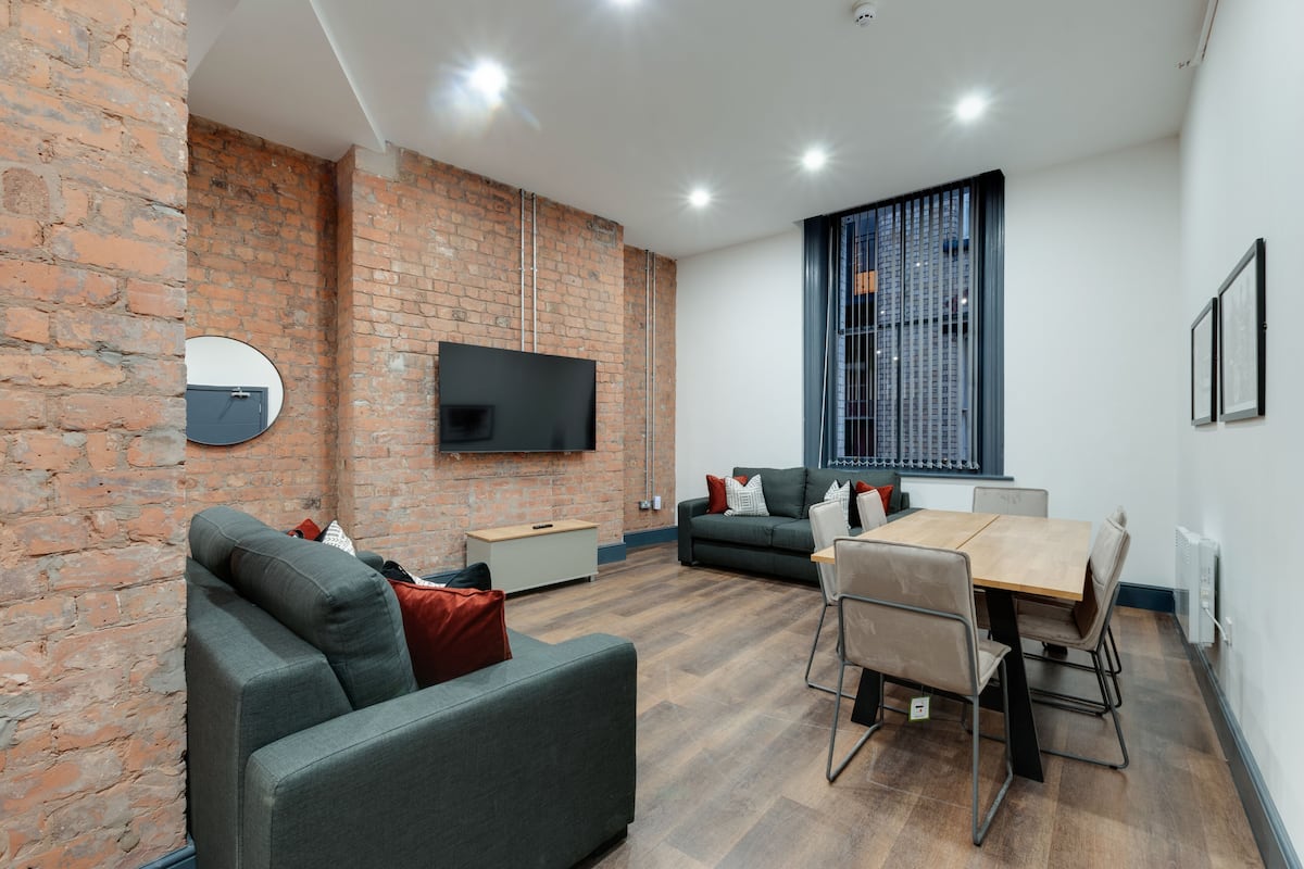 A modern living room with a brick wall, a flat-screen TV mounted above a sofa, a dining table with chairs, and a large window with blinds.