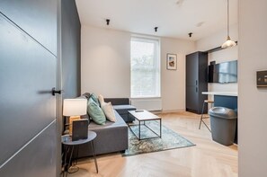 Cozy living space with a sofa, table, and lamp, featuring a kitchen area with a range hood and natural light.