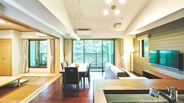 Habitació d'hotel moderna amb zona de dinar, un sofà, televisió i una finestra gran amb vistes a vegetació.