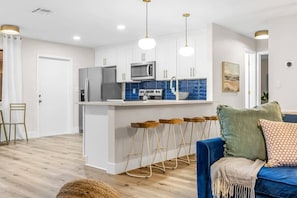 A modern kitchen with white cabinets, stainless steel appliances, and a blue backsplash. There are wooden stools at the island and a blue sofa with cushions.