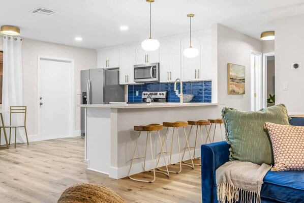 A modern kitchen with white cabinets, stainless steel appliances, and a blue backsplash. There are wooden stools at the island and a blue sofa with cushions.