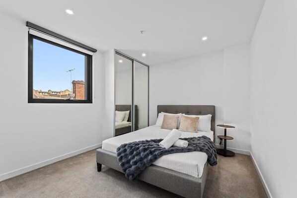 A modern bedroom with a large window, a bed with a grey headboard, and a small bedside table.
