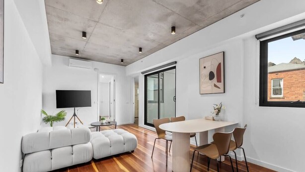 A modern living room with a sofa, a dining table, and chairs. There is a flat-screen TV mounted on the wall.