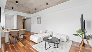 A modern living room with a sectional sofa, a dining area with chairs, a round coffee table, and a television on a tripod stand.