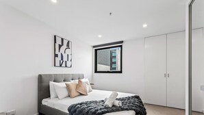 A modern bedroom with a grey bed, white walls, a window with a view, and a wardrobe.