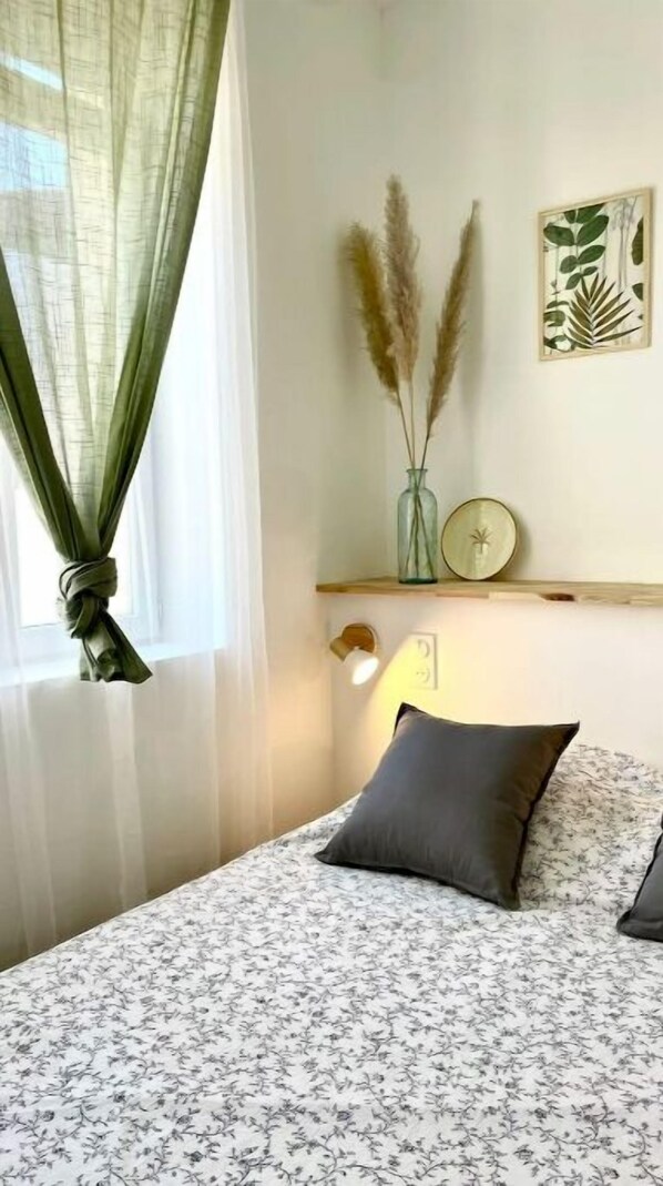 A bedroom with a bed, a bedside lamp, a window with sheer curtains, and a wall-mounted shelf with decorative items.