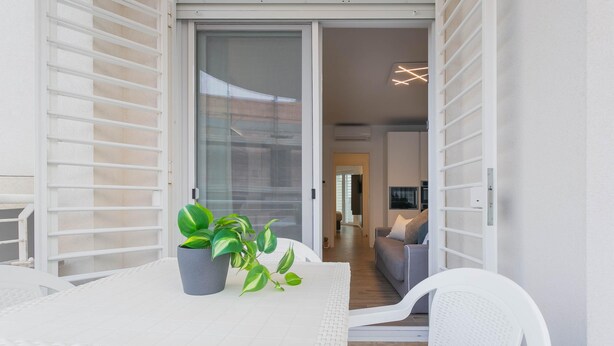 Ein modernes Wohnzimmer mit einer Schiebeglastür, die auf einen Balkon führt, einem weißen Tisch mit Topfpflanze und einem weißen Stuhl.