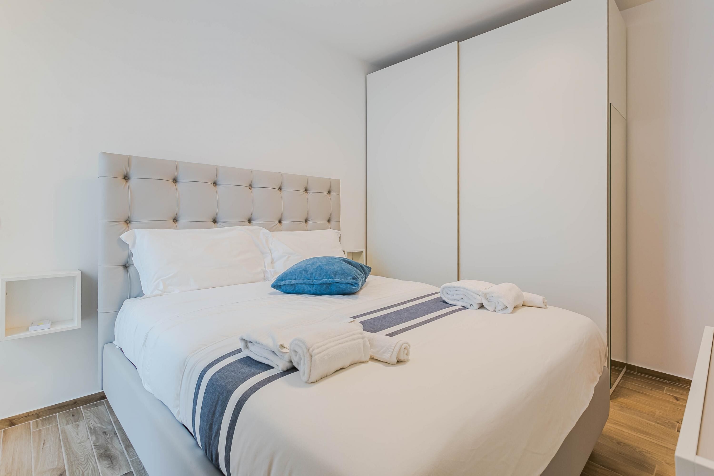 Una cama bien hecha con un cabecero capitonado, ropa de cama blanca, una almohada azul y toallas enrolladas.