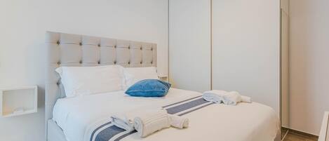 Una cama bien hecha con un cabecero capitonado, ropa de cama blanca, una almohada azul y toallas enrolladas.