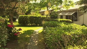 A garden pathway leading to a building with a tiled roof and a striped awning. - Villa Altea 2 Dans un Splendide Parc Fleuri, Calme à 900 Mètres de la Plage (Calvi)