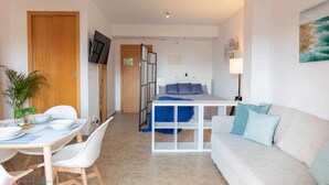 A modern hotel room with a bed, a sofa, a dining table with chairs, and a TV mounted on the wall.