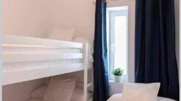 Habitació amb lliteres, una làmpada penjant, una finestra amb cortines i una petita planta al davant de la finestra.