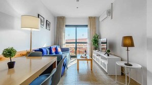 A modern living room with a sofa, a dining table, and a TV. There is a balcony visible through the window.