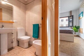 Un baño moderno con lavamanos, inodoro y bidé. Hay una toalla color turquesa colgada en la pared. La habitación tiene un ventanal con vistas a edificios.