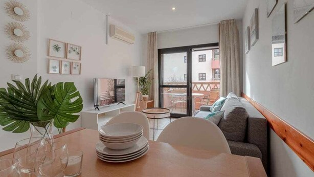 A modern living room with a dining area, a sofa, and a TV. There are plants, a window, and a balcony visible.
