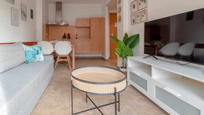A modern living room with a sofa, a round rattan table, a TV on a white stand, and a kitchen in the background.
