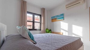 A bedroom with a bed, a window, an air conditioner, and a painting on the wall.