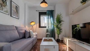 Ein modernes Wohnzimmer mit einem grauen Sofa, einem weißen Couchtisch, einer Stehlampe, einem Fernseher und einer großen Pflanze.