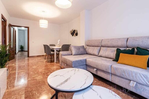 A modern living room with a grey sofa, two round marble tables, and a dining area with chairs.