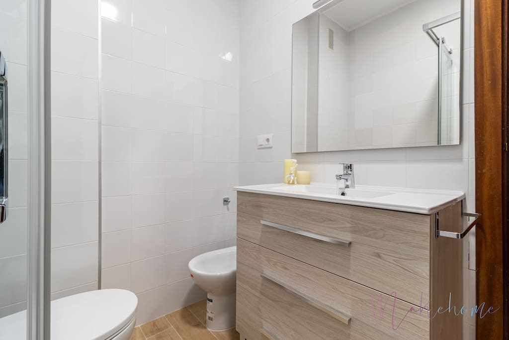 Un baño moderno con azulejos blancos, cabina de ducha de vidrio, inodoro blanco y tocador de madera con espejo.