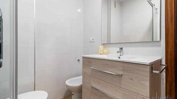 Un baño moderno con azulejos blancos, cabina de ducha de vidrio, inodoro blanco y tocador de madera con espejo.