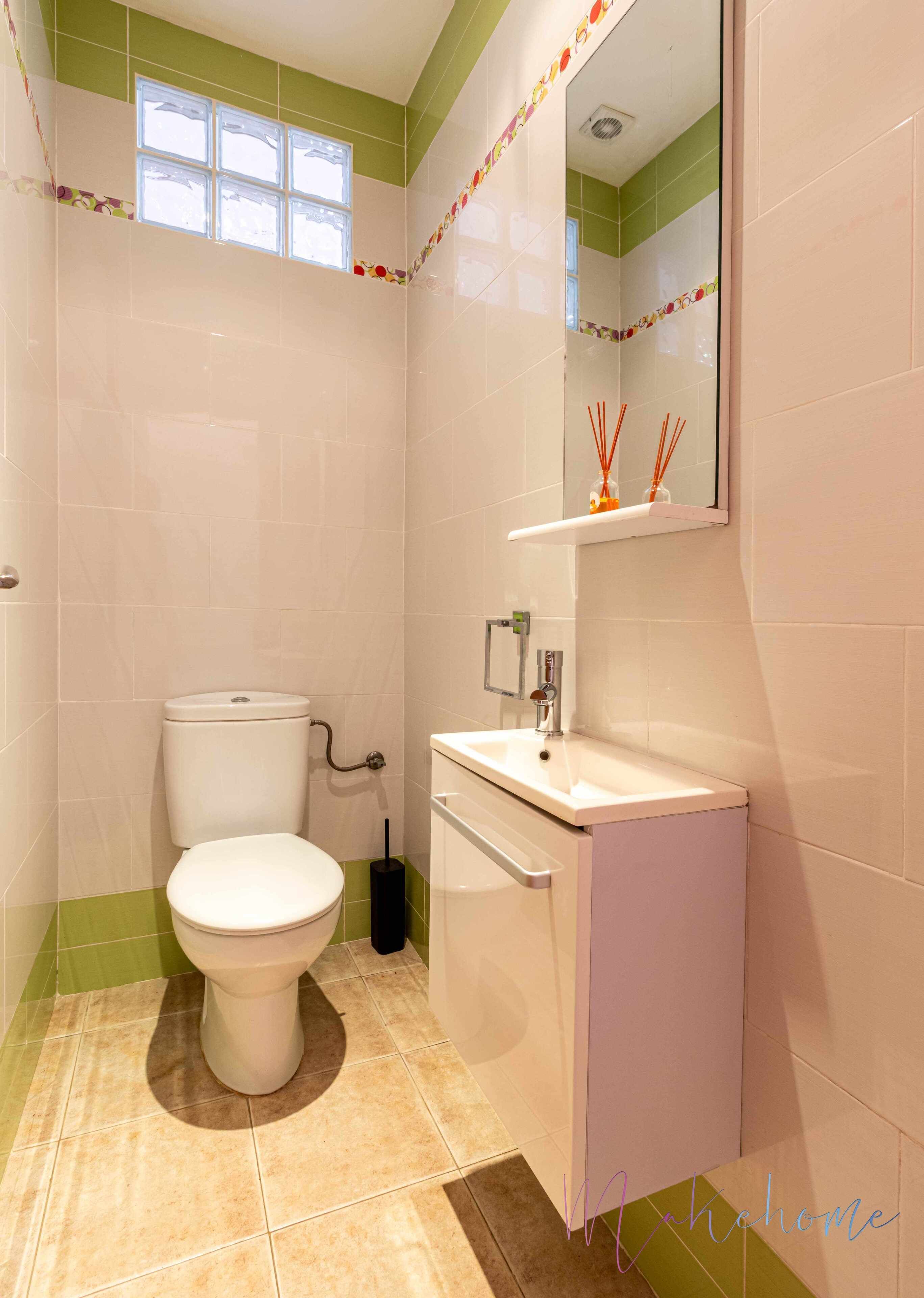 A bathroom with a toilet, sink, and mirror. There is a window above the toilet and a shelf with decorative items above the mirror.