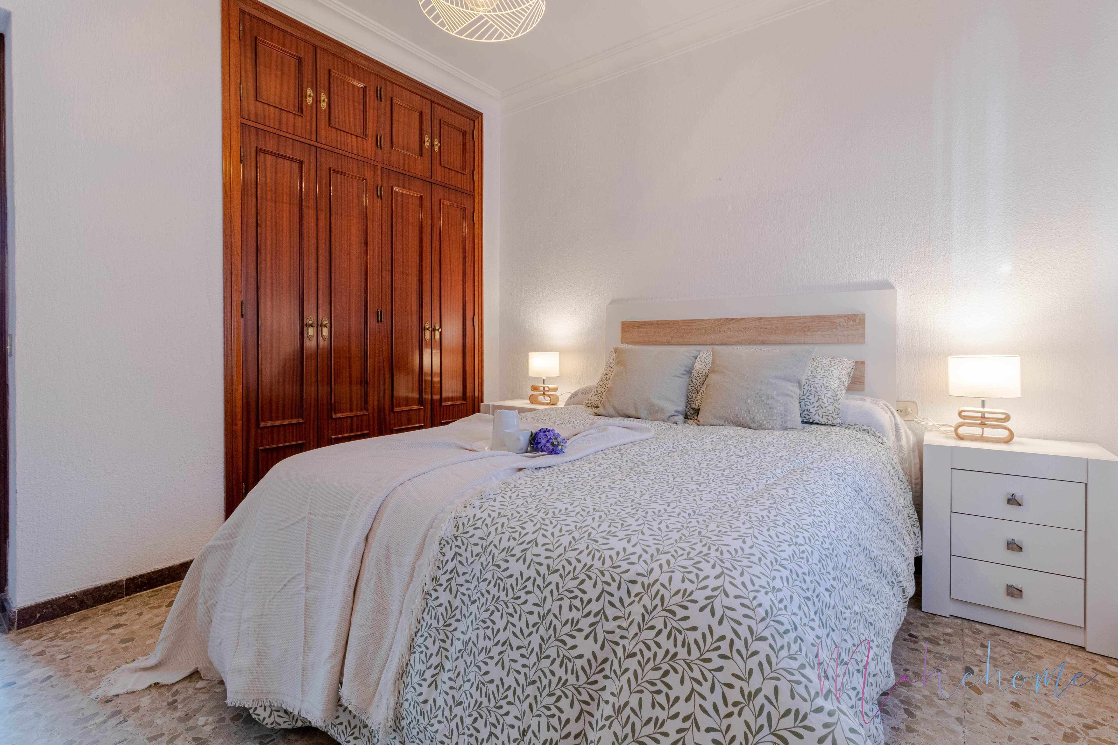 Une chambre à coucher avec une grande armoire en bois, un lit recouvert d’une couvre-lit à motifs, deux tables de chevet avec des lampes et une petite table de chevet ornée d’un objet décoratif.