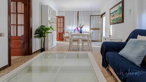 A modern living room with a glass coffee table, a sofa, a dining table with chairs, and a large window with sheer curtains. - Chalet a pie de Playa (Punta Umbria)