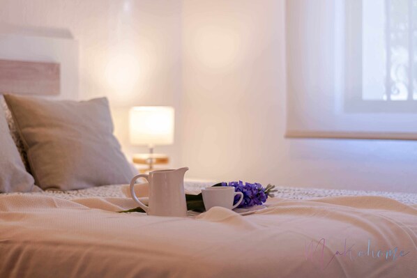 A neatly made bed with a white comforter, a white pitcher, a white cup, and a purple flower on the bed.