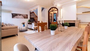 A spacious living room with a large wooden dining table, a sofa, and a small coffee table. There is a staircase leading to an upper floor, and a fireplace in the background.
