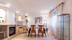 A dining area with a fireplace, a wooden table, and chairs. There are framed pictures on the wall and a window with curtains.