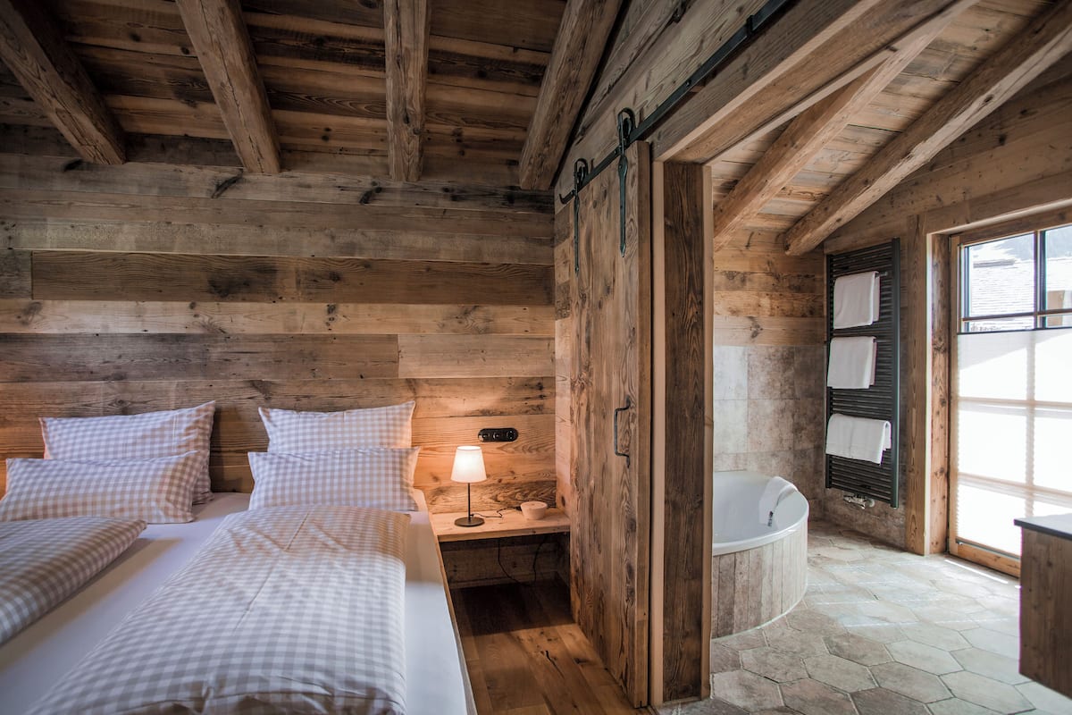 A wooden cabin interior with two beds, a nightstand with a lamp, a bathtub, and a window with towels hanging.