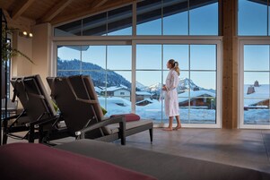 Ein Wellnessbereich mit Korbstühlen und einem großen Fenster mit Blick auf einen verschneiten Berg.