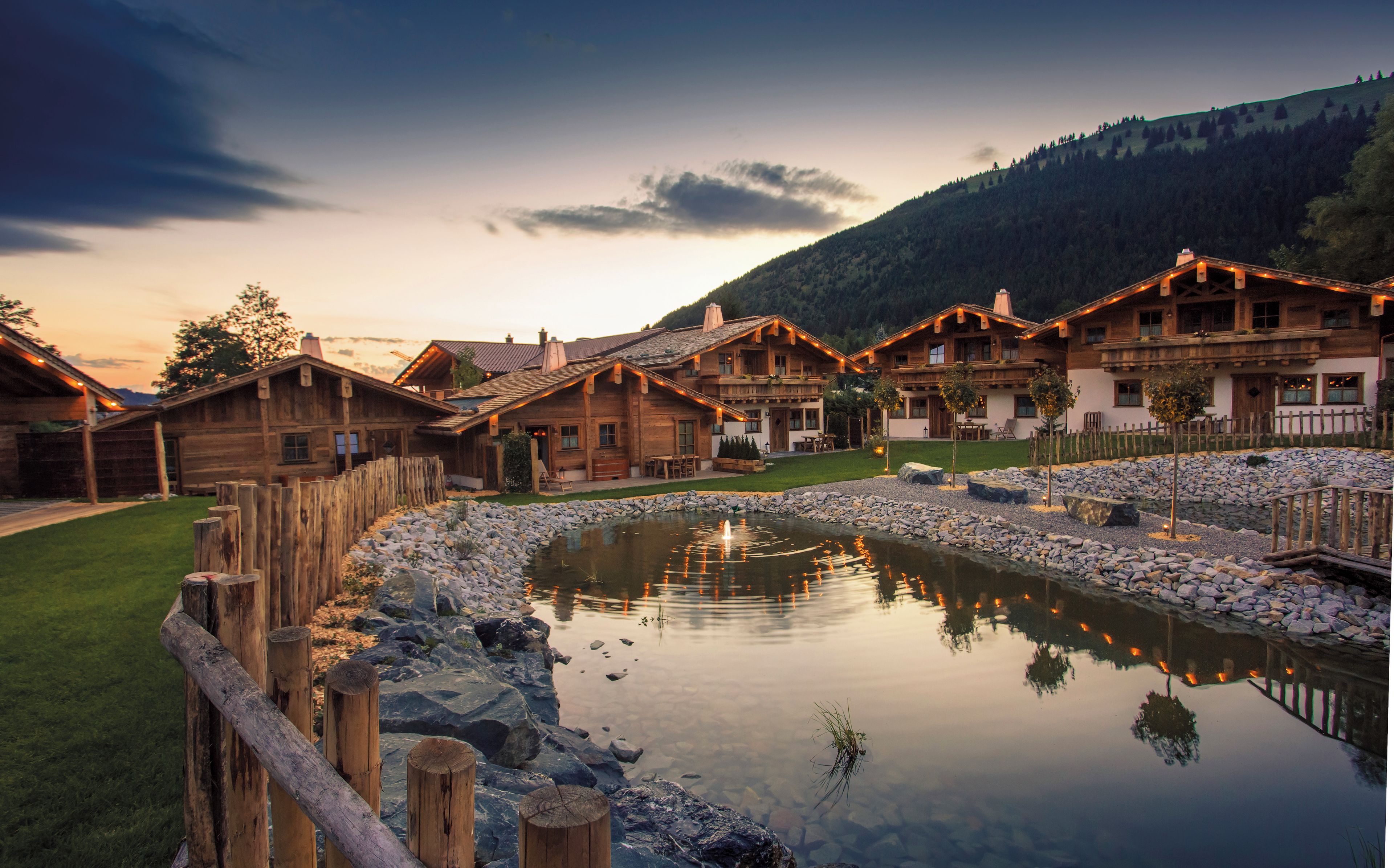Eine Holzlodge mit einem Teich, umgeben von einem Holenzaun, vor einer Bergkulisse.