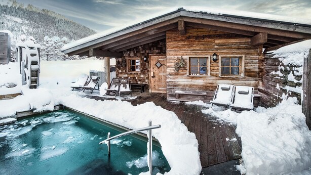 A wooden cabin with a pool surrounded by snow, wooden deck, and lounge chairs. - Alpin Chalets Oberjoch (Bad Hindelang)