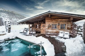 A wooden cabin with a pool surrounded by snow, wooden deck, and lounge chairs. - Alpin Chalets Oberjoch (Bad Hindelang)