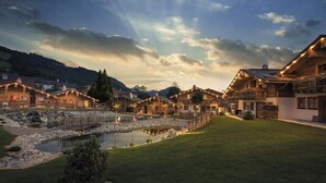 A traditional chalet-style building with a stone pathway leading to a wooden bridge over a small pond, surrounded by lush greenery and a mountainous backdrop. - Alpin Chalets Oberjoch (Bad Hindelang)