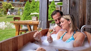 A wooden hot tub with two people holding glasses of white wine, surrounded by a wooden deck and a view of a chalet-style building. - Alpin Chalets Oberjoch (Bad Hindelang)
