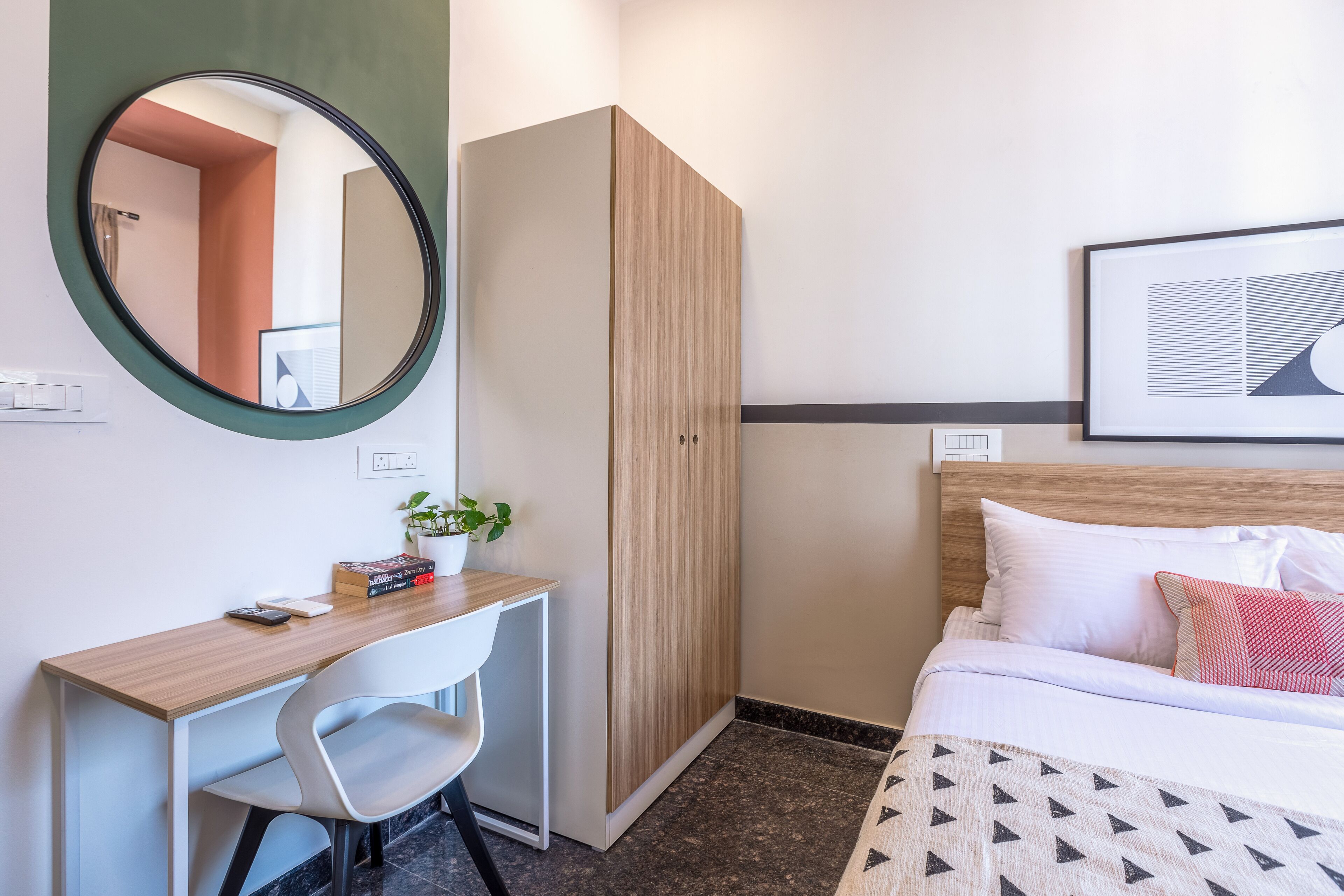 Een hotelkamer met een bed, een bureau met stoel, een garderobe, een ronde spiegel en een lijst met een afbeelding aan de muur.