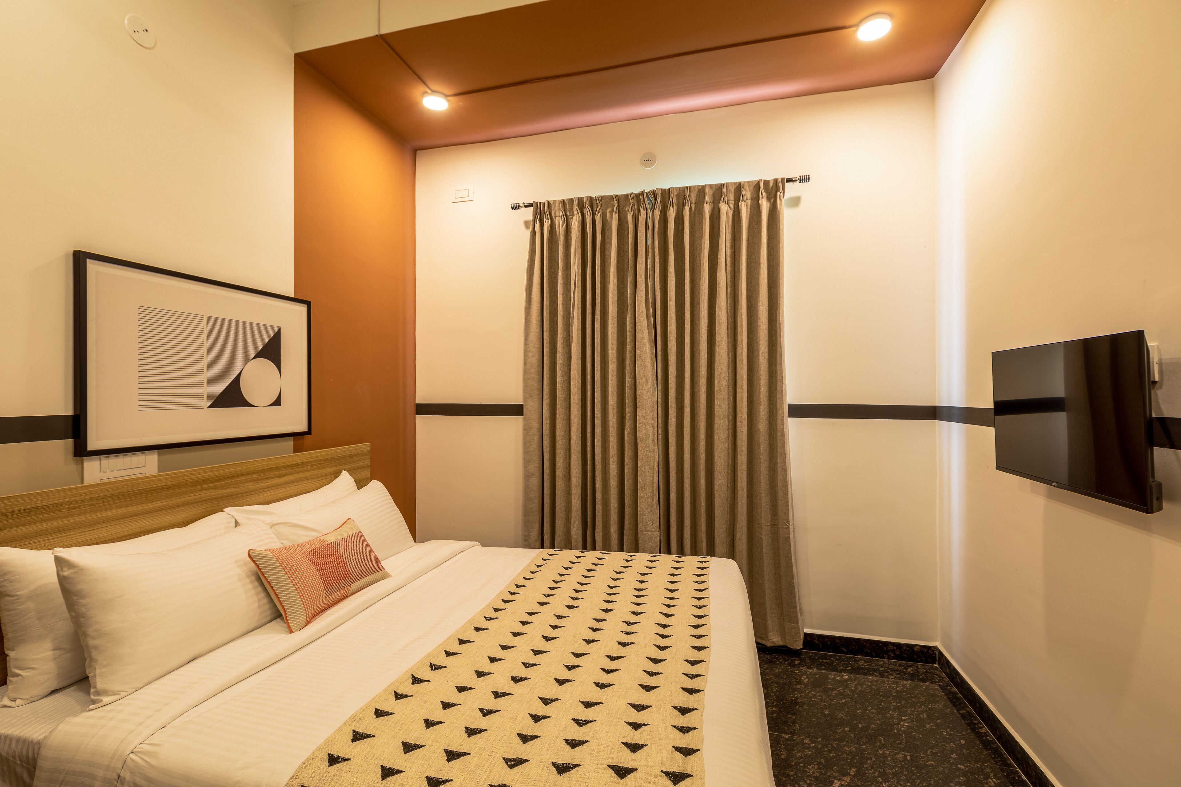 A hotel room with a bed, a TV mounted on the wall, a framed picture, and a window with curtains.