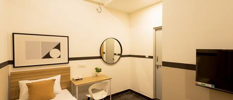 A hotel room with a bed, a desk with a chair, a round mirror, a TV, and an air conditioning unit.