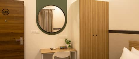 A hotel room with a wooden door, a desk with a chair, a bed with pillows, a round mirror, and a wardrobe.