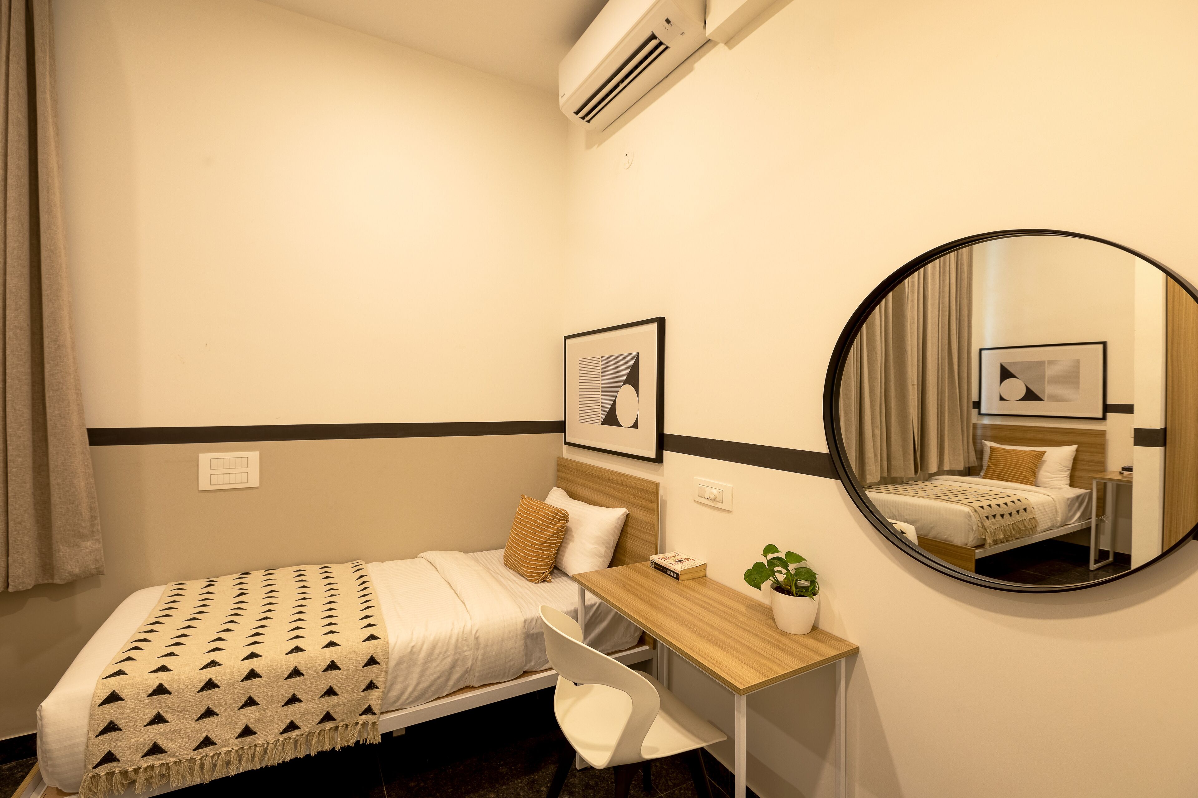 Hotelkamer met een bed, bureau, stoel, ronde spiegel, potplant en een lijst aan de muur.