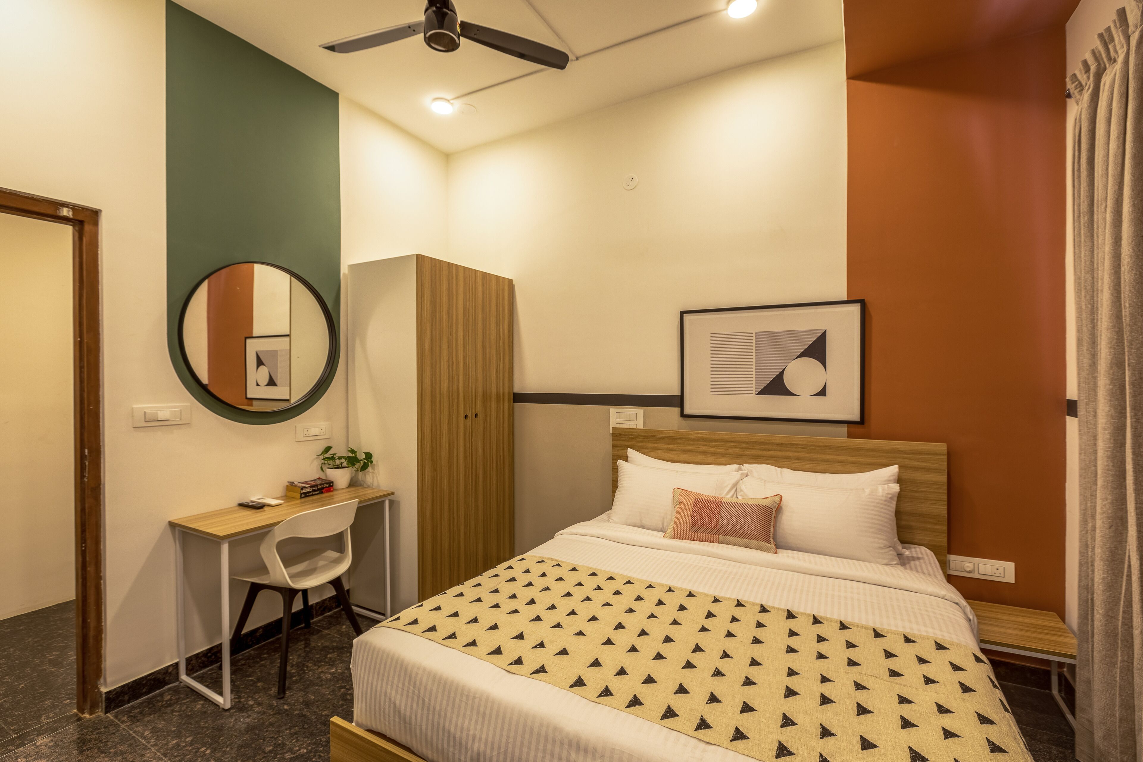 A neatly arranged hotel room with a bed, a desk, a chair, a wardrobe, and a ceiling fan.