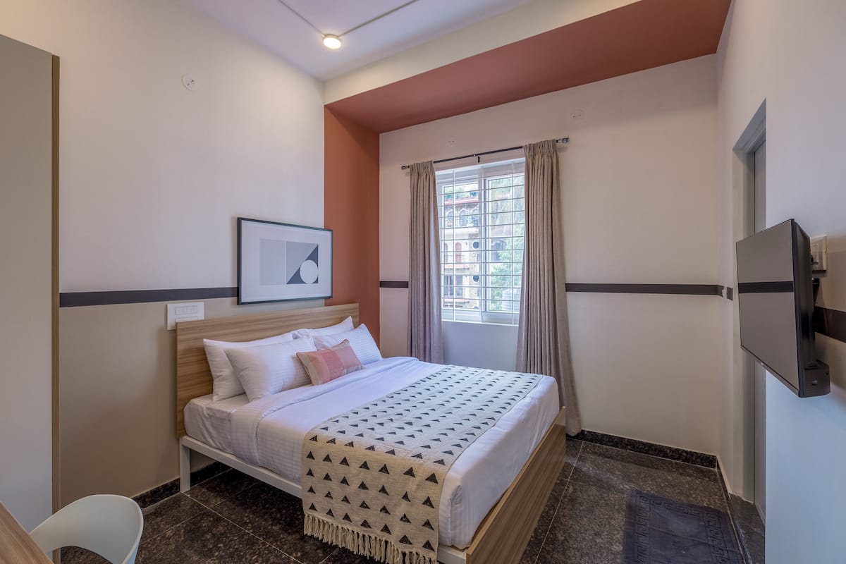 A hotel room with a bed, a TV mounted on the wall, a window with curtains, and a framed picture on the wall.